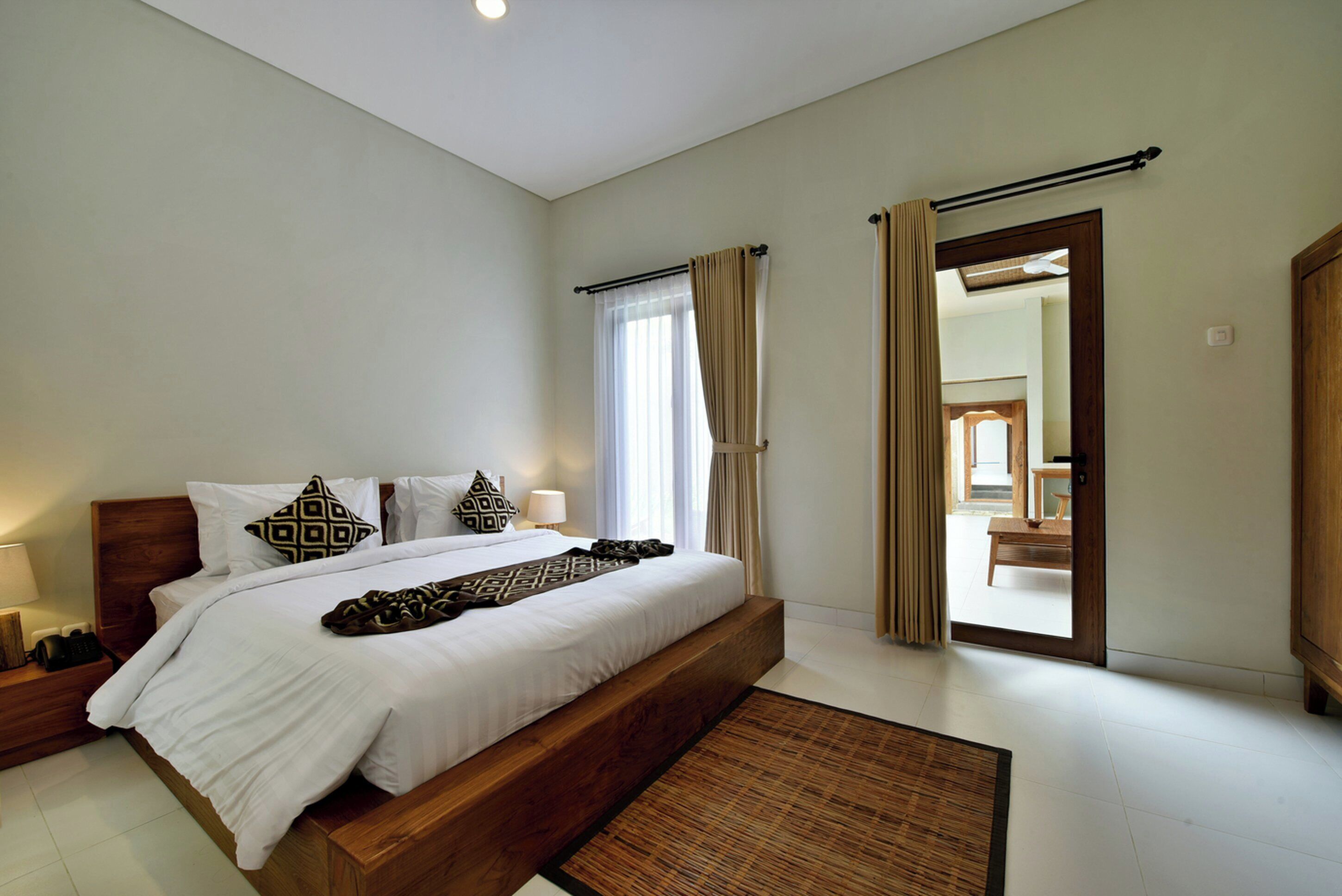 Une chambre avec un grand lit, des tables de chevet, une commode, un banc en bois, une fenêtre avec des rideaux et une porte donnant sur une autre pièce.
