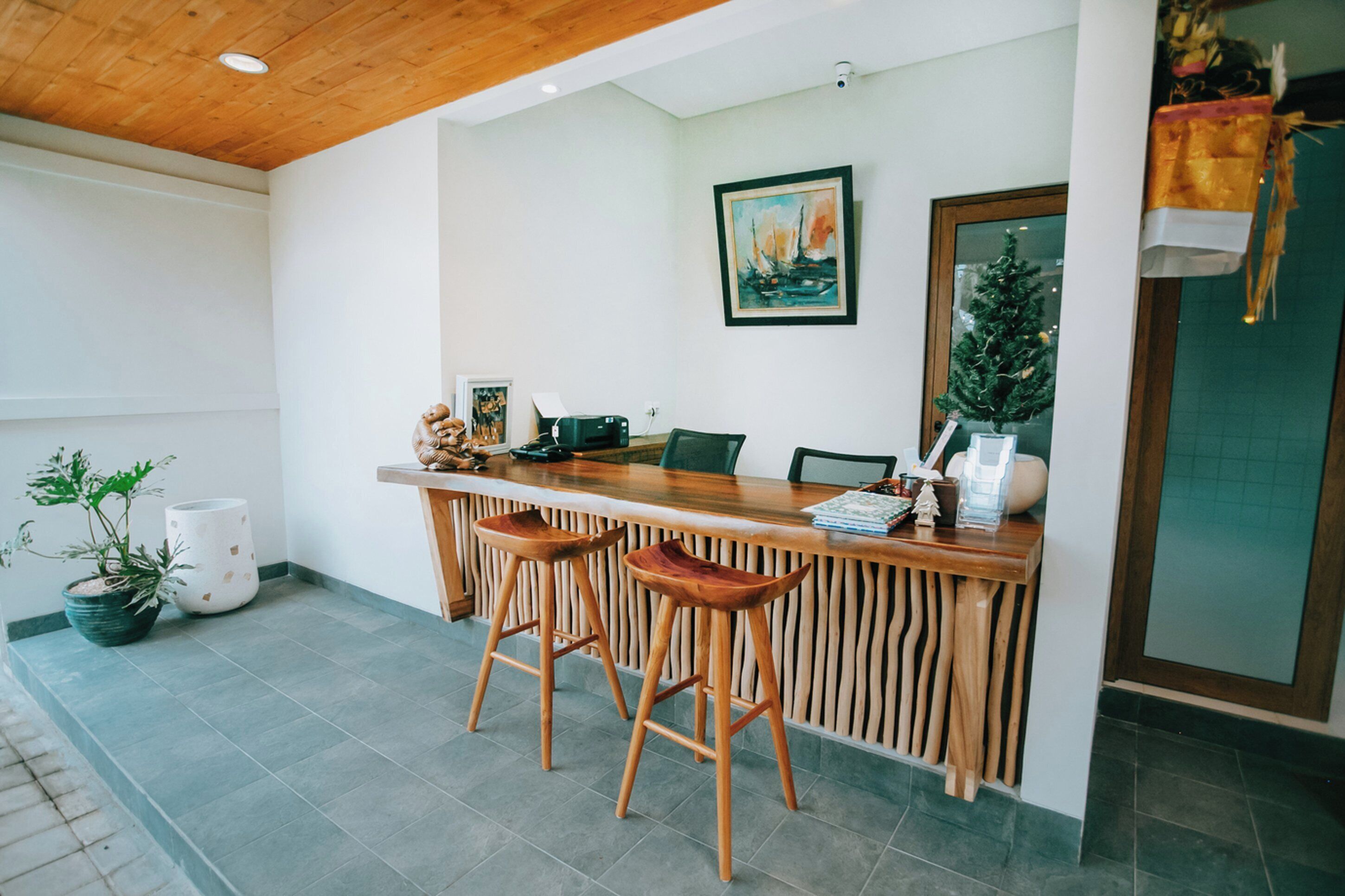 A reception area with a wooden reception desk, two wooden barstools, a potted plant, a framed painting, and a small Christmas tree.