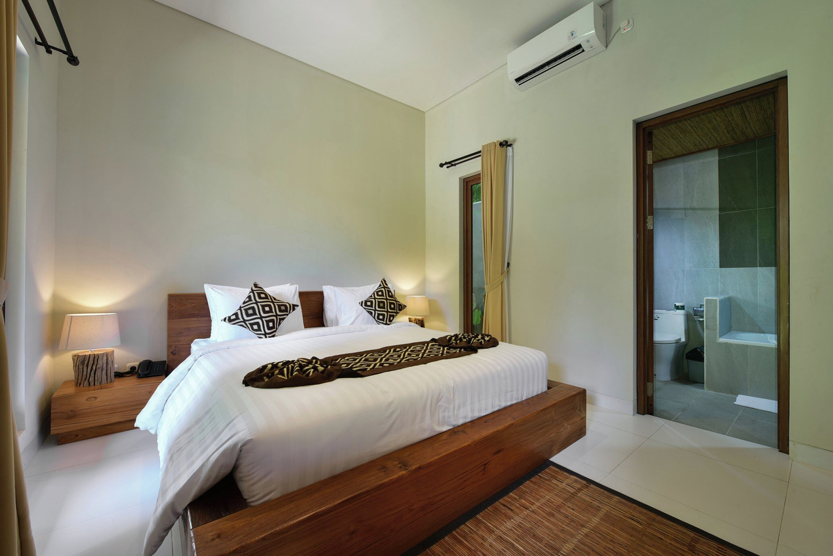 Une chambre d’hôtel moderne avec un grand lit, une tête de lit en bois, une table de chevet avec une lampe et une porte-fenêtre coulissante donnant sur la salle de bain.