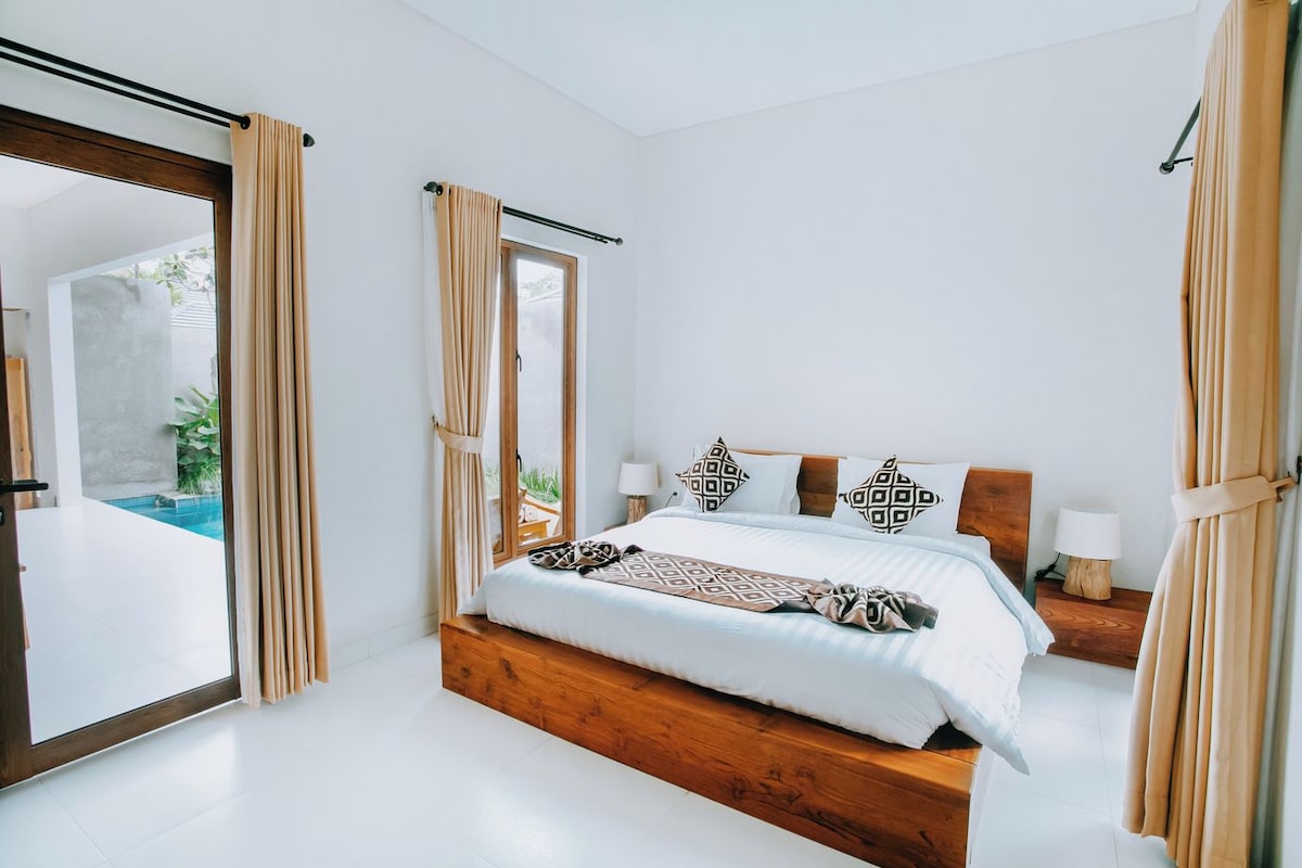 A bedroom with a large bed, a wooden headboard, two bedside tables with lamps, and a view of an outdoor swimming pool.