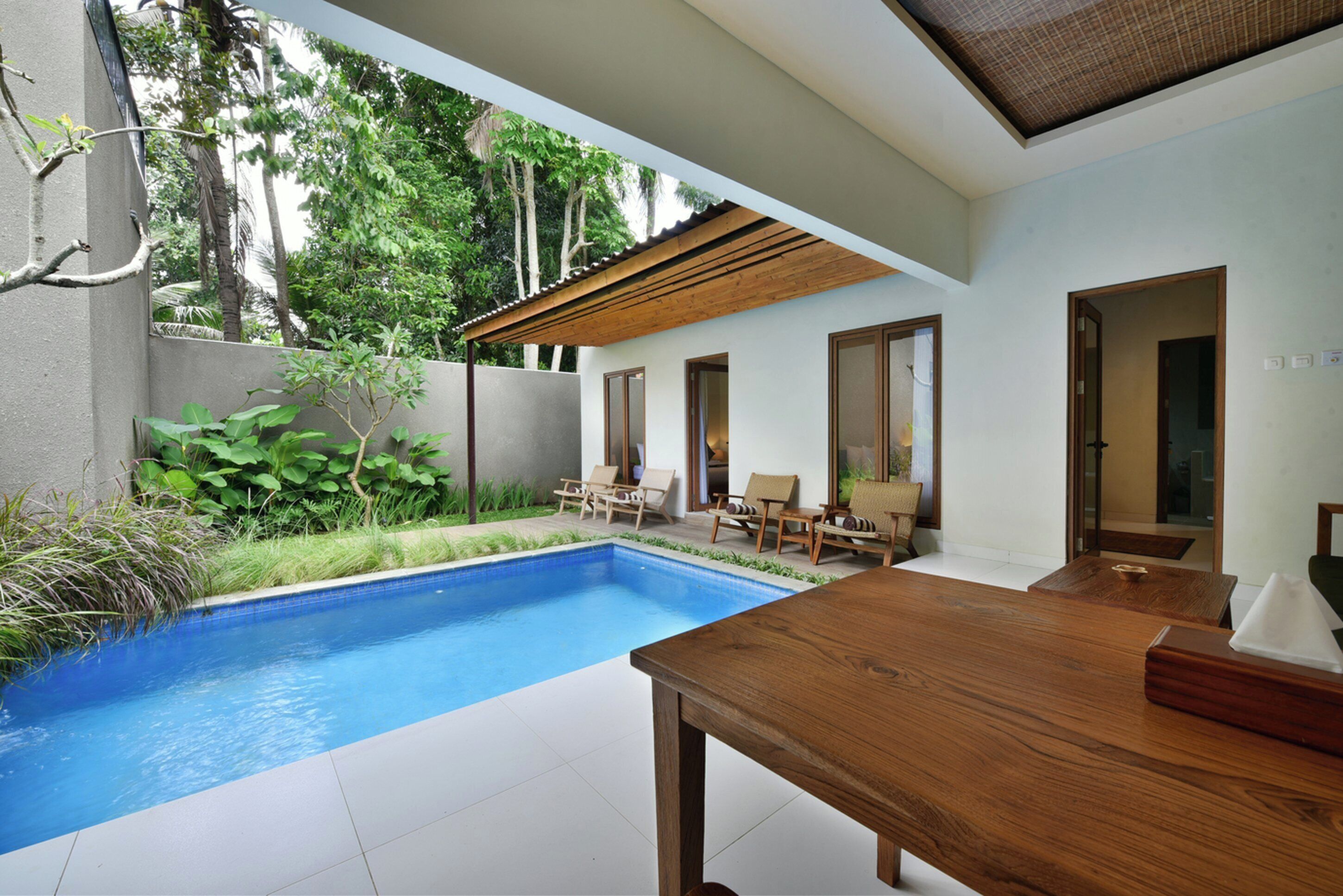 Un espace piscine moderne avec une table et des chaises en bois, entouré de verdure et bordé d’un mur blanc.