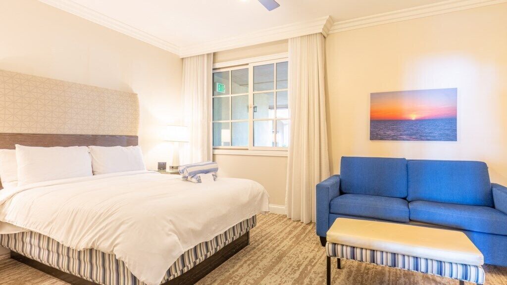 A hotel room with a bed, a blue sofa, a bench, a painting, a window, and a lamp.
