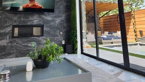 Een moderne woonkamer met een grote televisie bevestigd aan een donkere marmeren muur, een glazen salontafel met een plant, en uitzicht op de omgeving via grote schuifpuien.