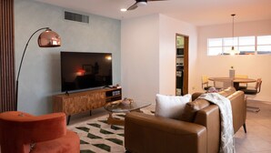 Un salon moderne doté d’un téléviseur à écran plat, d’un ventilateur de plafond, d’un coin repas avec une table et des chaises, et d’une cuisine visible à travers une porte ouverte.