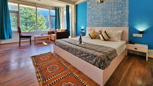 Un dormitorio amplio con una cama grande, una mesita de noche, un escritorio y una silla. Hay ventanas grandes con cortinas, un papel tapiz estampado y un suelo de madera.