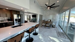 A modern kitchen with a large island, bar stools, and a dining area with a table and chairs. - Sea U Soon - Luxury Dock Home (Fort Myers Beach)