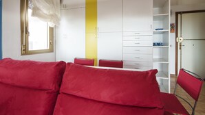 A room with a red sofa, a yellow vertical accent wall, white cabinets, and a small dining area with a red chair. - Verde Mare in Punta Pedrosa (Golfo Aranci)