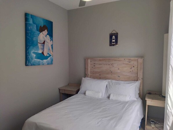 A bedroom with a wooden headboard, white bedding, a nightstand, a small wall-mounted shelf, and a painting of a woman in a swimsuit. - 5 Telena (eMdloti)