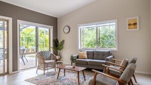 Un salon spacieux avec un canapé gris, des fauteuils en osier, une table basse et de grandes fenêtres donnant sur un paysage verdoyant.
