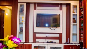 A living room with a TV mounted on a wooden cabinet, surrounded by shelves with decorative items and a small table with a vase of flowers. - Sacred Trails Homestay (Burhanilkantha)