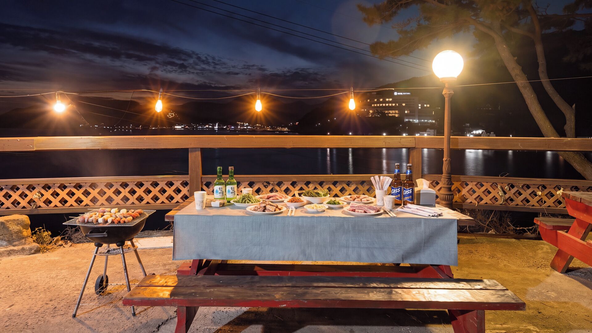 An outdoor dining area with a table set for a meal, overlooking a body of water with city lights in the background.