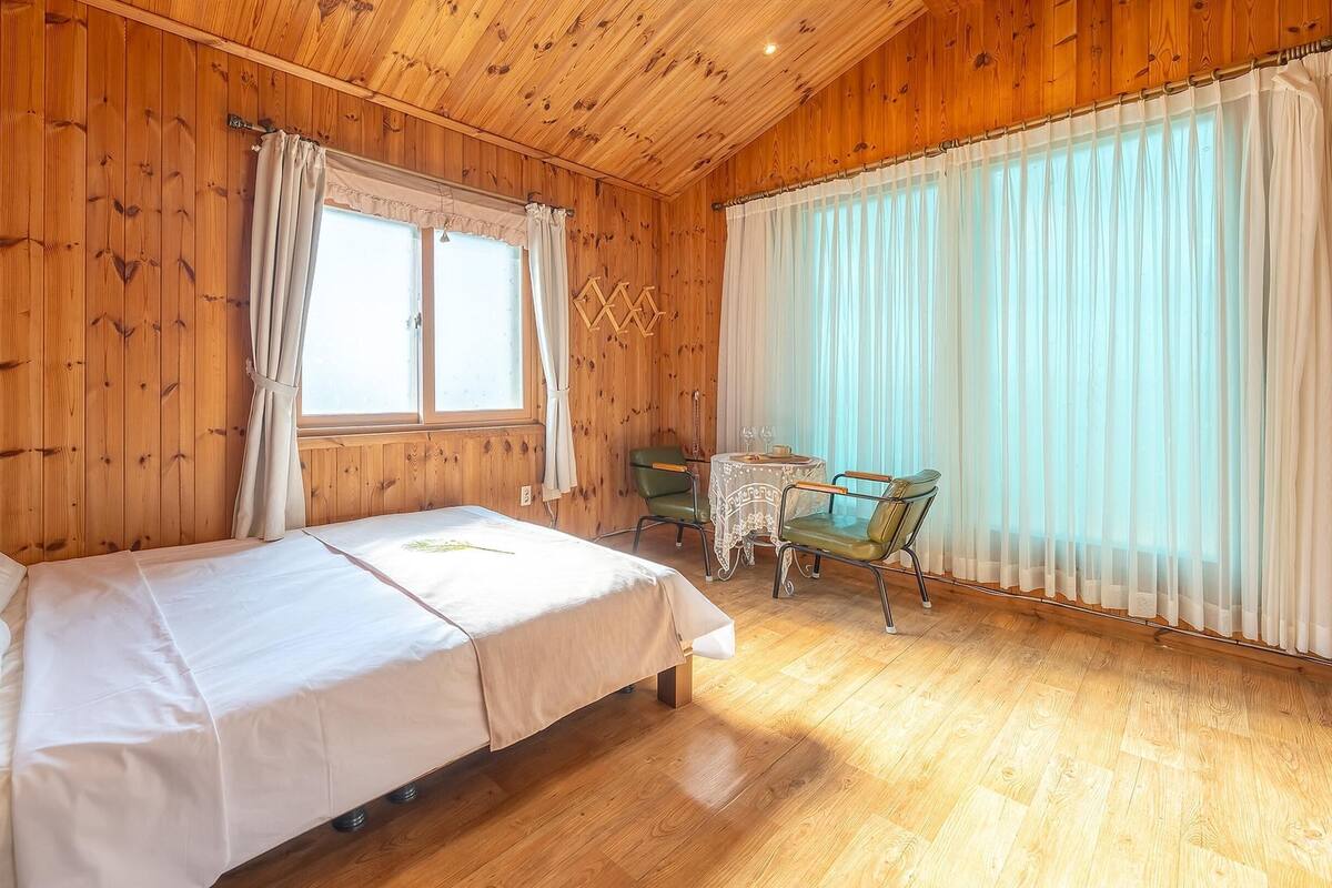 A wooden-paneled room with a bed, a table with chairs, and large windows with sheer curtains.