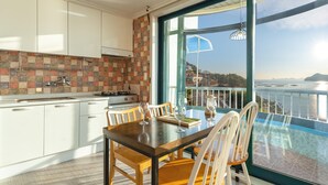 Una cocina moderna con armarios blancos, salpicadero de azulejos y un comedor con mesa y sillas. Un ventanal ofrece vistas al mar y una isla en la distancia.