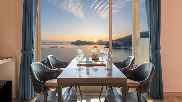 Una mesa de comedor con cuatro sillas, una botella de agua y dos copas de vino dispuestas para dos personas. La mesa está frente a un gran ventanal con vistas a un atardecer sobre el agua y colinas al fondo.