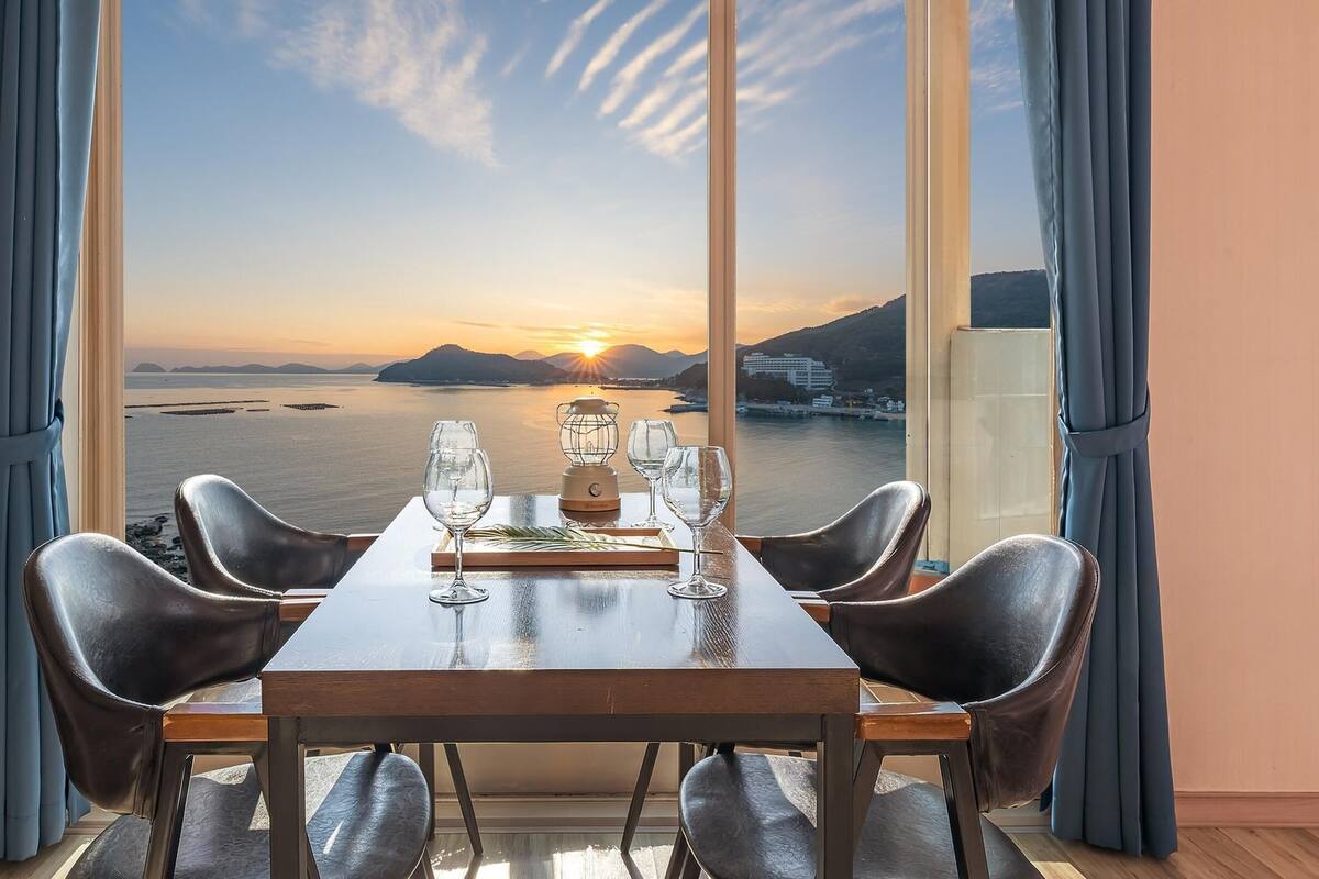 A dining table with four chairs, a bottle of water, and two wine glasses set for two. The table is positioned in front of a large window with a view of a sunset over a body of water and distant hills.