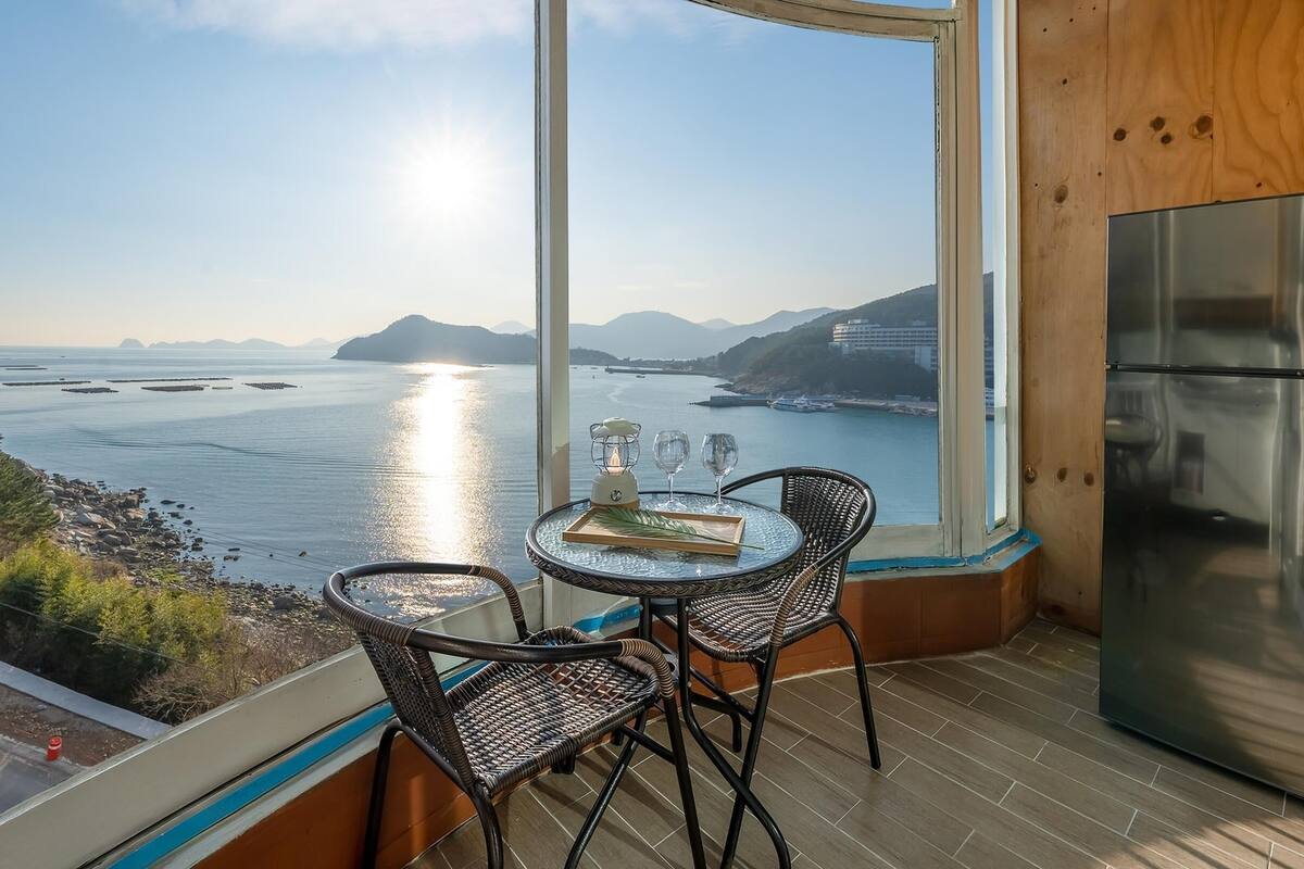 A balcony with a view of the sea, a table with two chairs, and a refrigerator.
