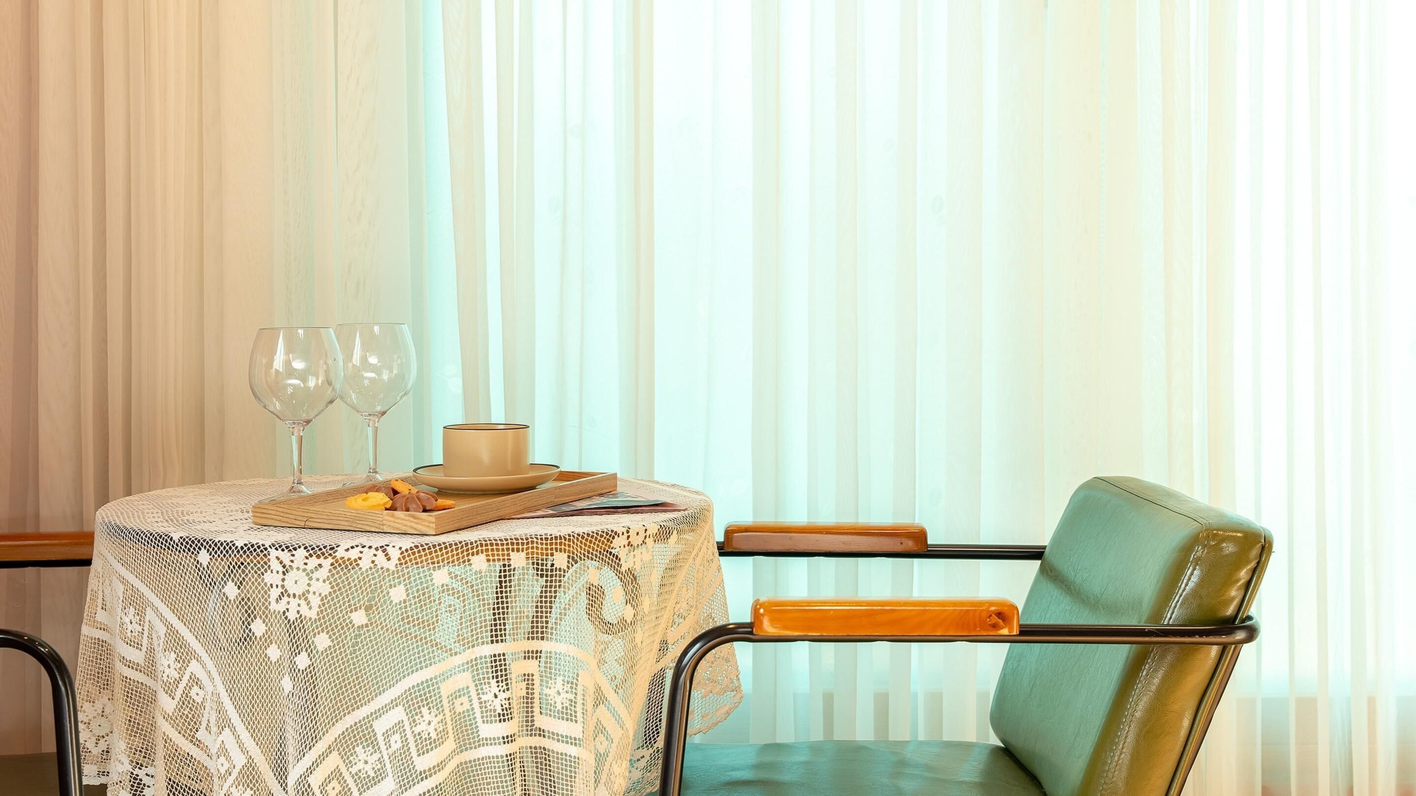 Une table recouverte d’une nappe en dentelle, avec deux verres à vin, une tasse et une assiette remplie de collations. Une chaise verte est placée à côté de la table.