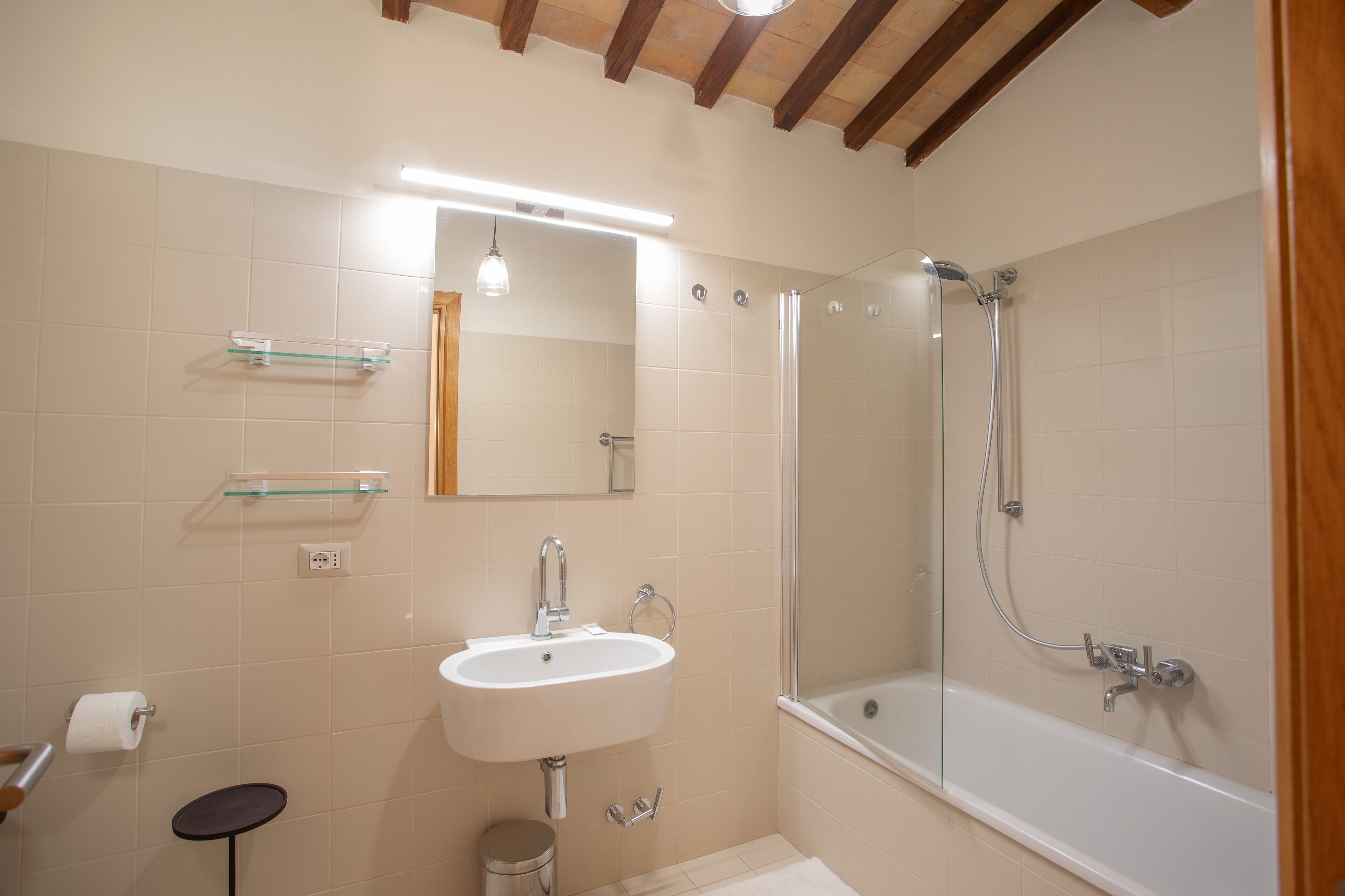 Un baño con un lavamanos blanco, estantes de vidrio, un espejo, una ducha y un techo de madera.