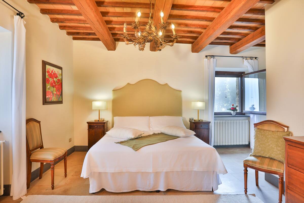 Un dormitorio con techo de madera, una cama grande con ropa blanca, dos mesitas de noche con lámparas, una silla y una ventana con vista a árboles.