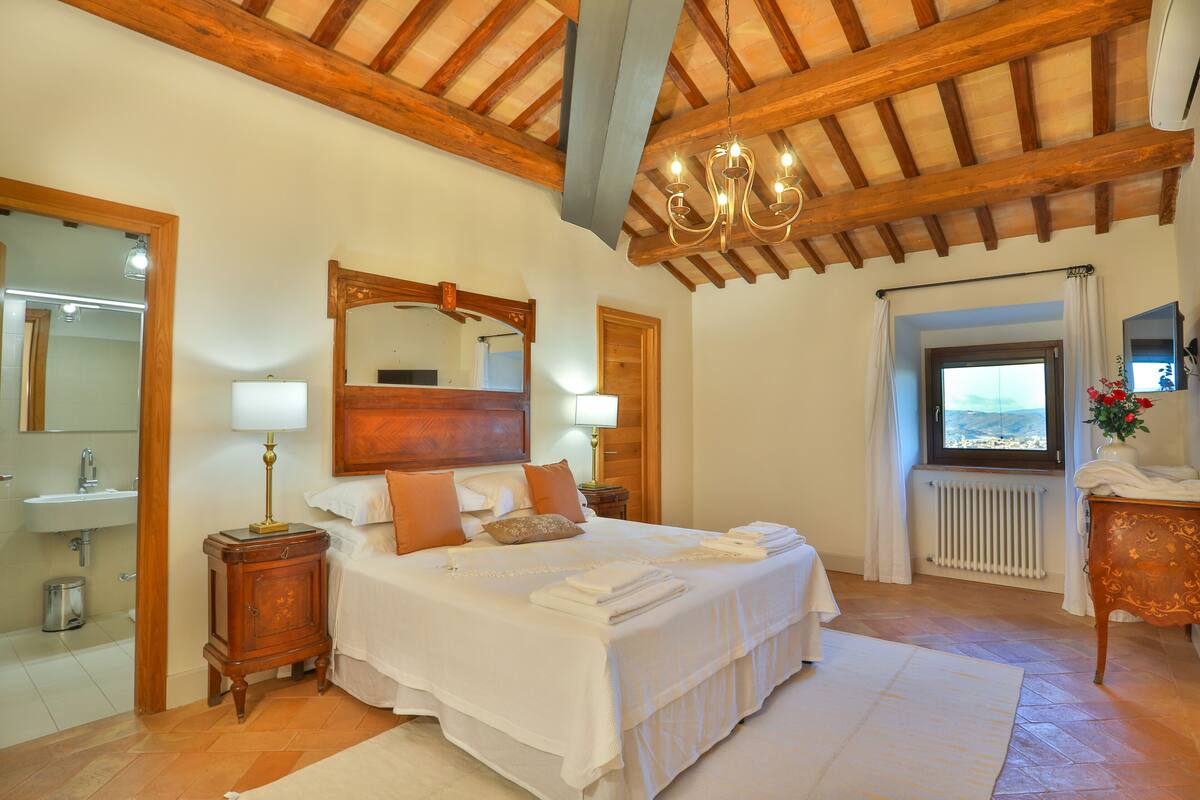 Un dormitorio con techo de madera, una cama grande con ropa blanca, una mesita de noche con lámpara, un aparador de madera y una ventana con vistas.