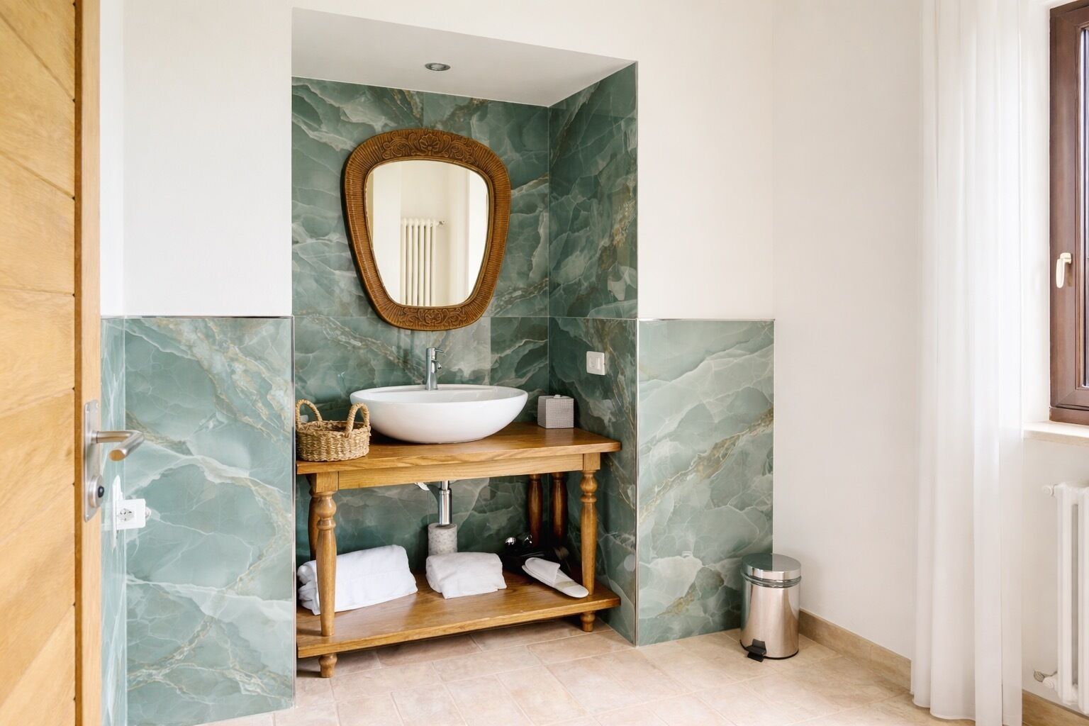 Un baño con pared de mármol, tocador de madera, lavamanos blanco, espejo y un bote de basura.