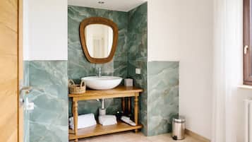 Un baño con pared de mármol, tocador de madera, lavamanos blanco, espejo y un bote de basura.