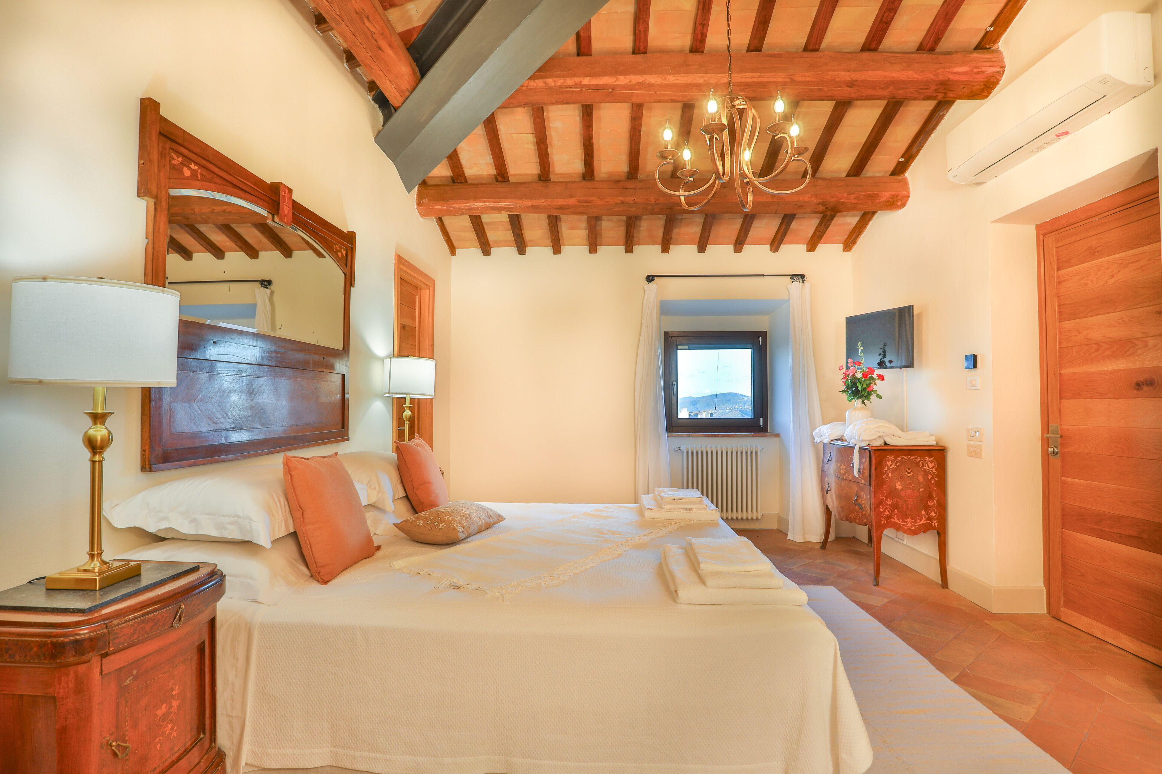 Une chambre avec un plafond en bois, un lit recouvert de draps blancs, une table de chevet, un miroir et une fenêtre donnant sur un beau paysage.
