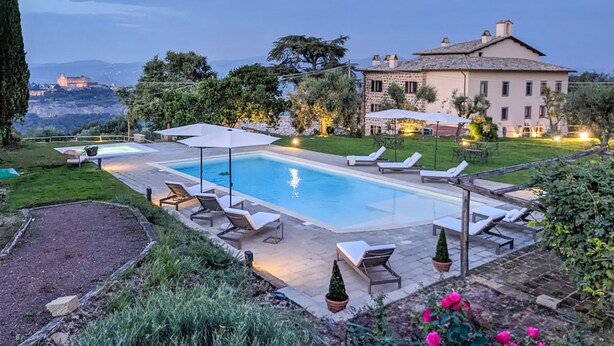 Zona de piscina con sillones de descanso y sombrillas, rodeada por un jardín y un edificio grande con fachada de piedra.
