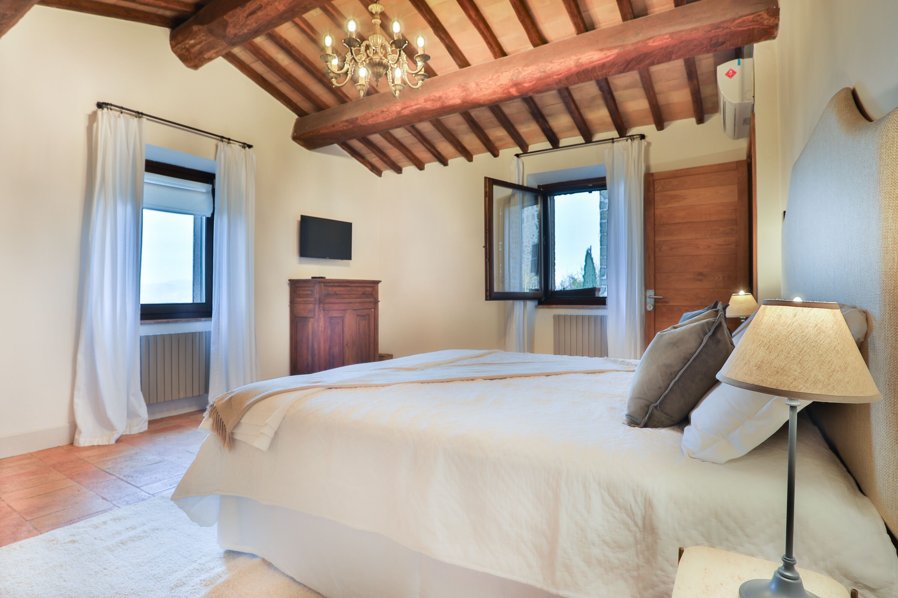 A bedroom with a large bed, wooden ceiling beams, a chandelier, a nightstand, and a window with curtains.