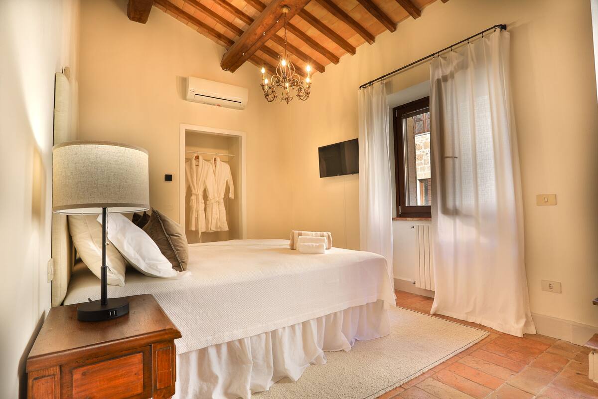 Un dormitorio con cama, lámpara de noche, armario, candelabro, ventana con cortinas y televisor de pantalla plana.