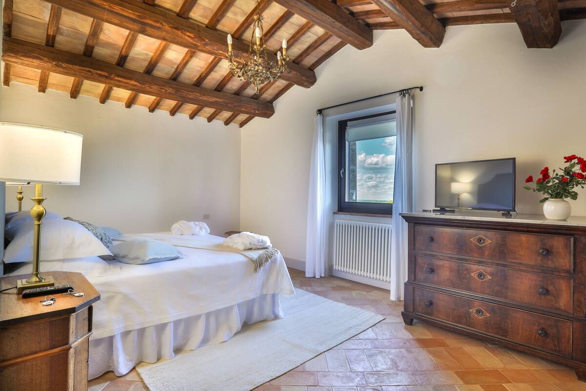 Un dormitorio con cama, cómoda, televisor y ventana con cortinas.