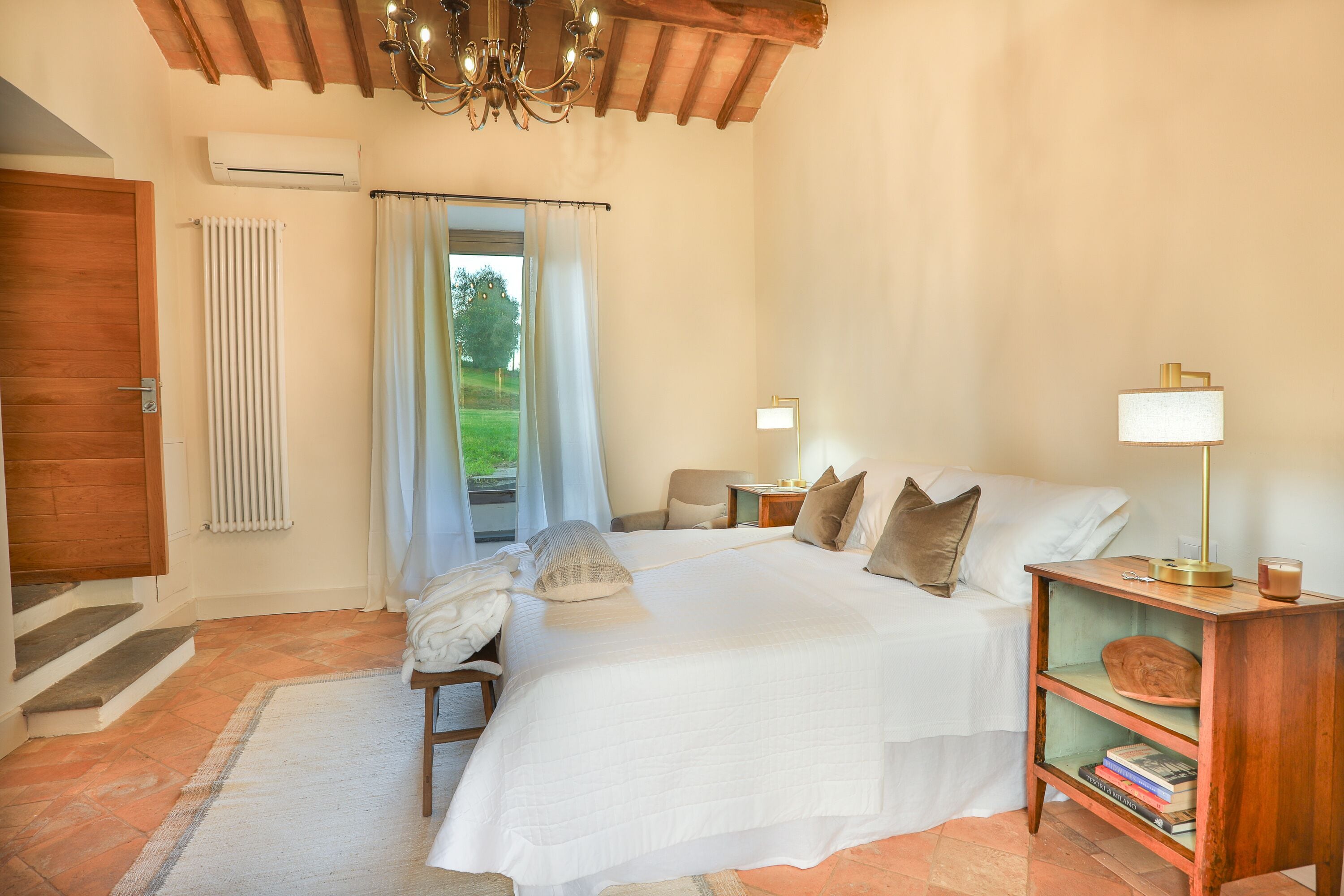 A bedroom with a large bed, a wooden nightstand, a lamp, a bookshelf, a chair, a window with curtains, and a radiator.
