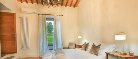 A bedroom with a large bed, a wooden nightstand, a lamp, a bookshelf, a chair, a window with curtains, and a radiator.
