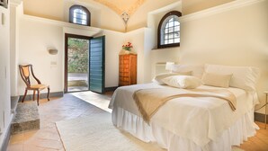 Un dormitorio con una cama grande, una silla de madera, un banco de piedra y vistas a un jardín por una puerta abierta.