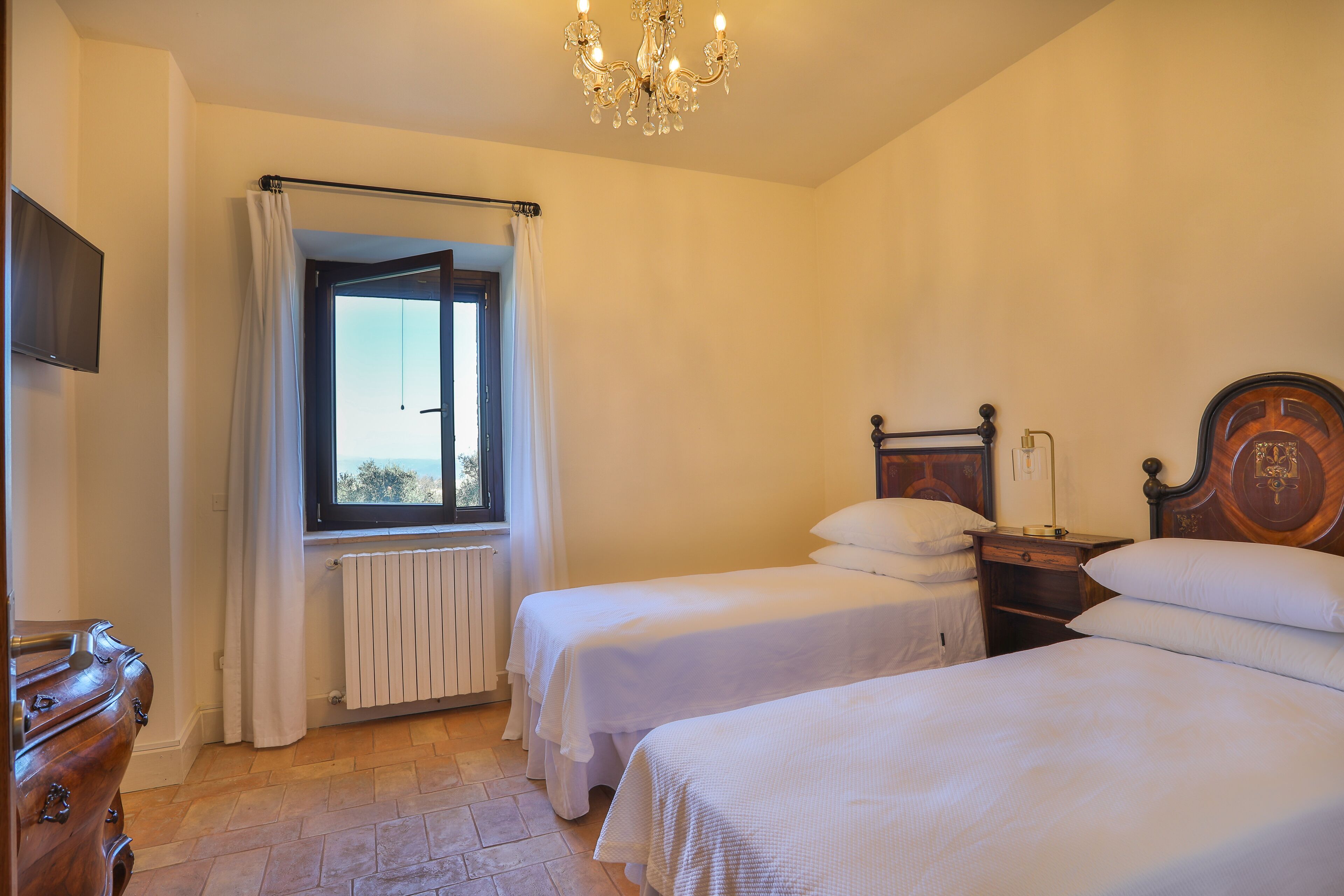 Een slaapkamer met twee eenpersoonsbedden, een houten dressoir, een radiator, een raam met gordijnen en een kroonluchter.
