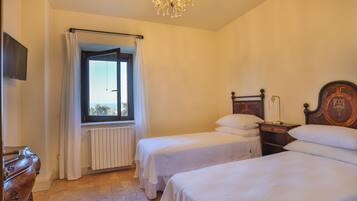 A bedroom with two single beds, a wooden dresser, a radiator, a window with curtains, and a chandelier.