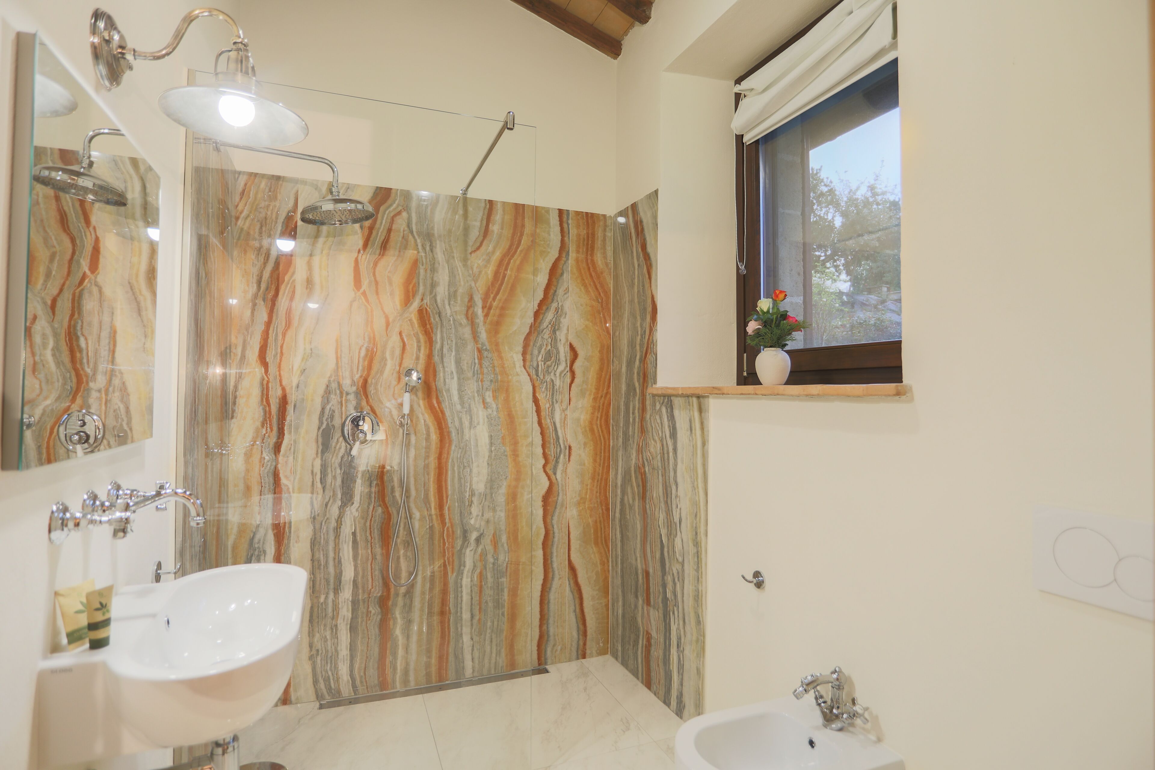 Une salle de bain avec un mur de douche en marbre, deux lavabos, un miroir et une fenêtre donnant sur la forêt.