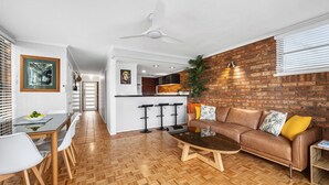 A modern living room with a herringbone wooden floor, a brown leather sofa with yellow cushions, a glass coffee table, and a white ceiling fan.