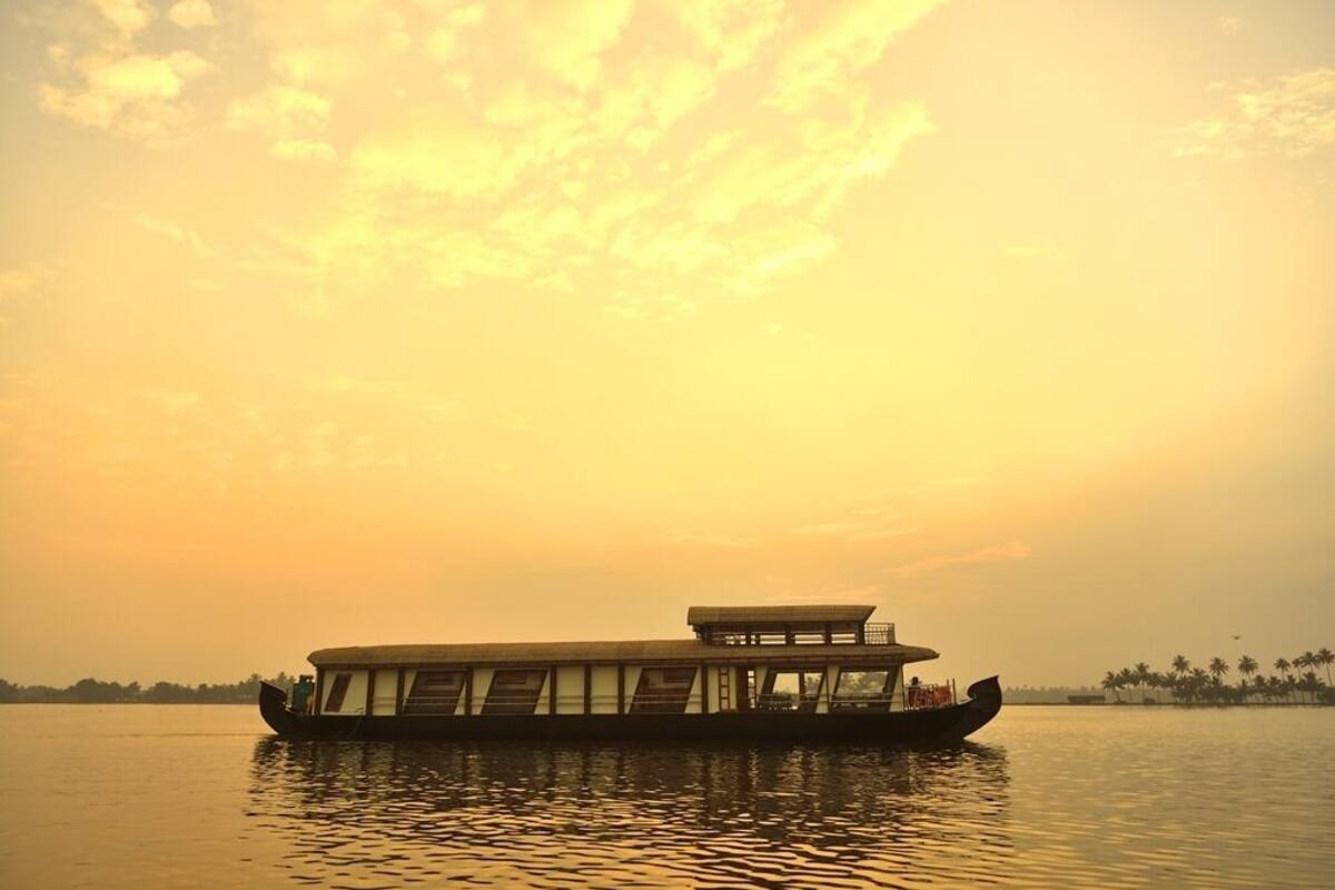 A riverboat with a covered deck and a small structure on top, floating on calm water during sunset.