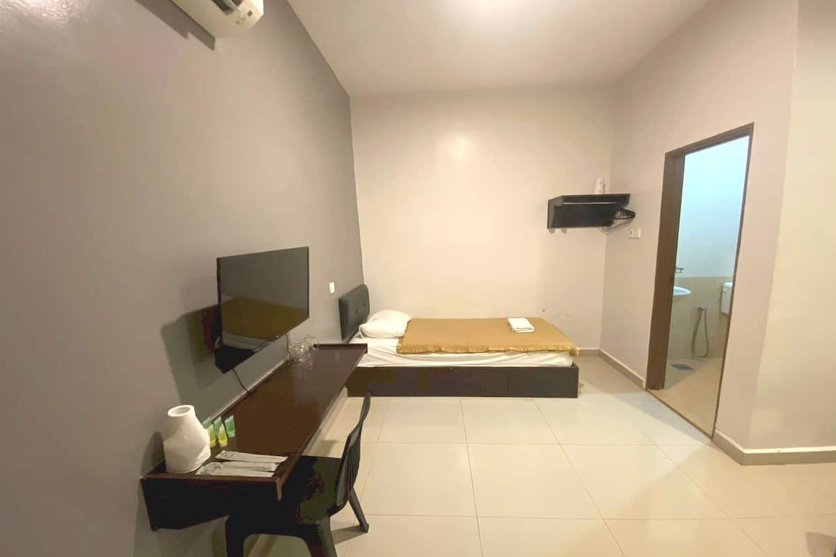 A small, modern bedroom with a bed, a desk with a chair, a television, and a bathroom visible through an open door.
