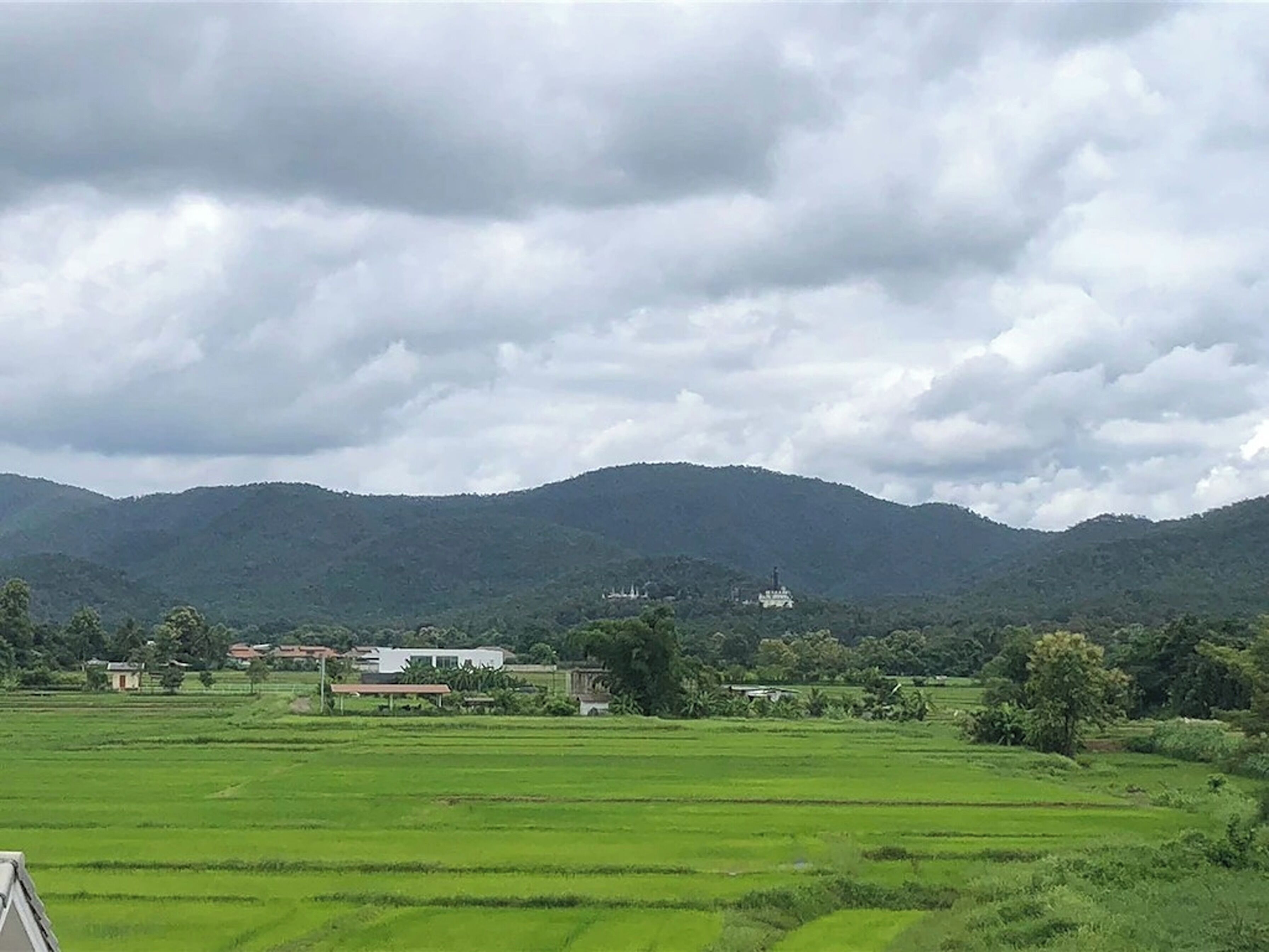 A rural landscape with green fields, a few buildings, and mountains in the background.