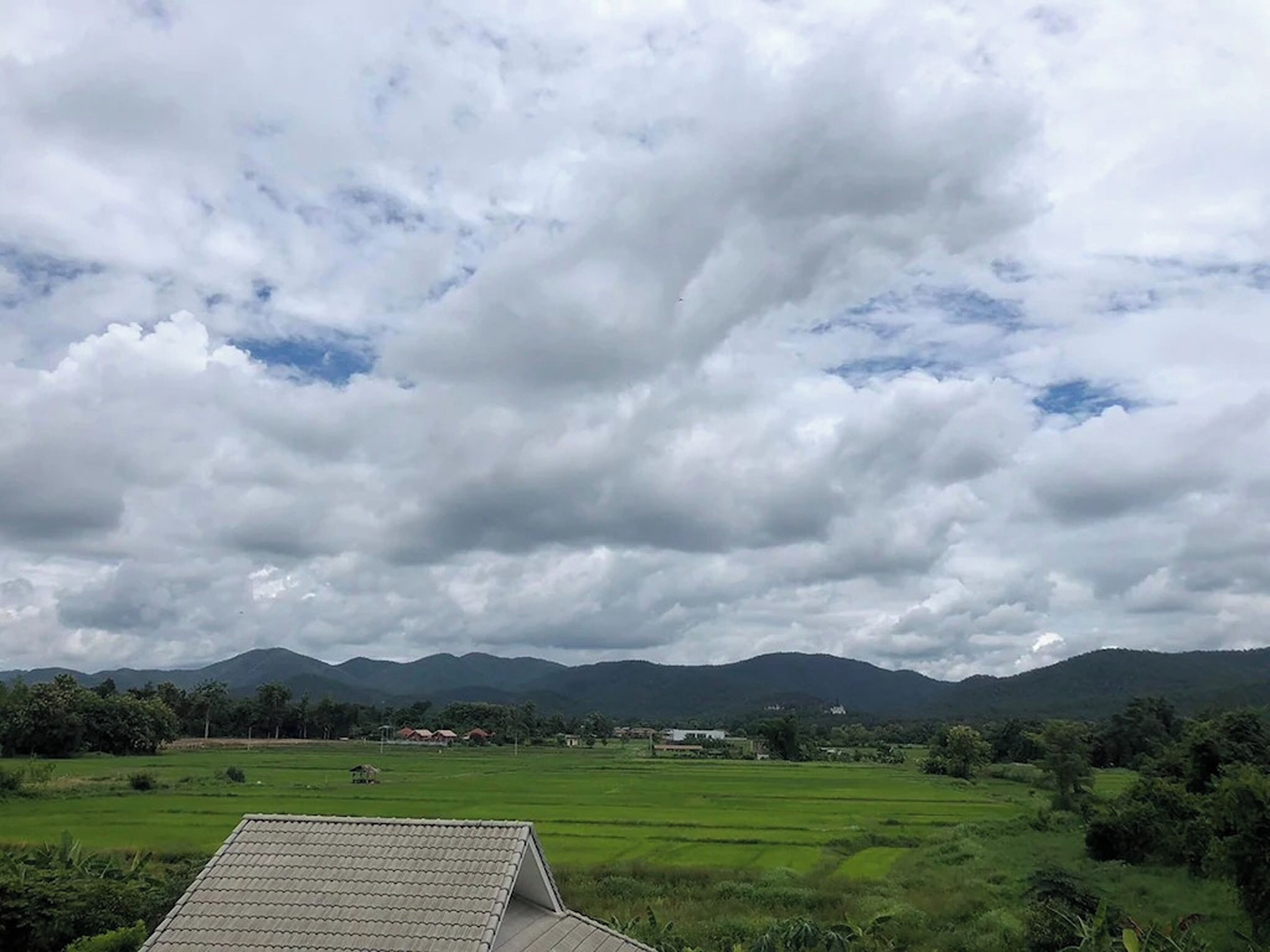 A rural landscape with green fields, a house with a tiled roof, and mountains in the background.