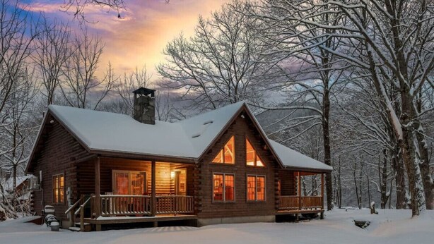 A log cabin with a porch, large windows, and a chimney, surrounded by snow-covered trees.