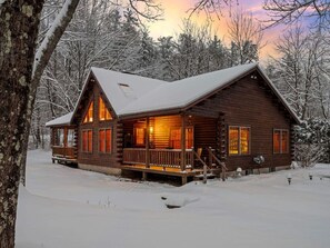 Una cabaña de troncos con un porche y ventanas amplias, rodeada de árboles cubiertos de nieve.
