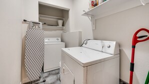 Een wasserette met een wasmachine, droger, strijkplank en schoonmaakspullen.