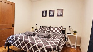 A bedroom with a patterned bedspread, a wooden door, a bedside table with a lamp, and framed botanical prints on the wall. - 3 Bedroom 10min Walk to the UofA/Hospital.   (Edmonton)