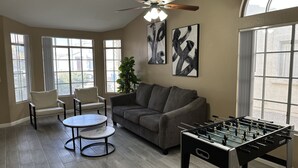 Een woonkamer met een grijze bank, twee fauteuils, een ronde salontafel en een tafelvoetbaltafel. Er zijn grote ramen met gordijnen, een plafondventilator en abstracte kunst aan de muren.