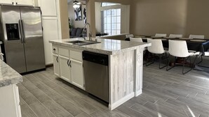 Een moderne keuken met een roestvrijstalen koelkast, witte kasten, een marmeren eiland en een eetgedeelte met witte stoelen en een zwarte tafelkleed.