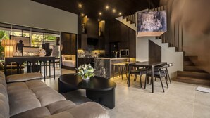 Una sala de estar moderna con sofá, mesa de centro y comedor. Se observa una cocina con barra y una escalera.