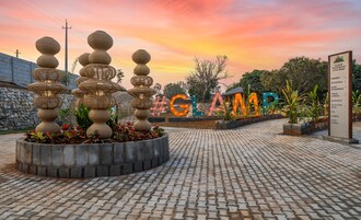 Área de acesso pavimentada com uma placa para Glamp Wilderness, apresentando um vaso de pedra com plantas e uma estrutura decorativa.